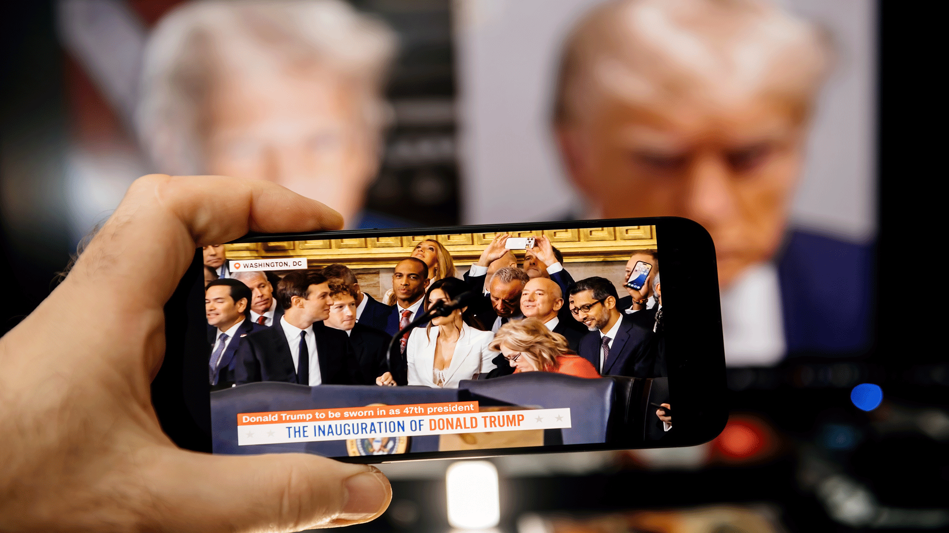 A hand holds a smartphone displaying a live broadcast of Donald Trump’s inauguration with Jeff Bezos and Sundar Pichai