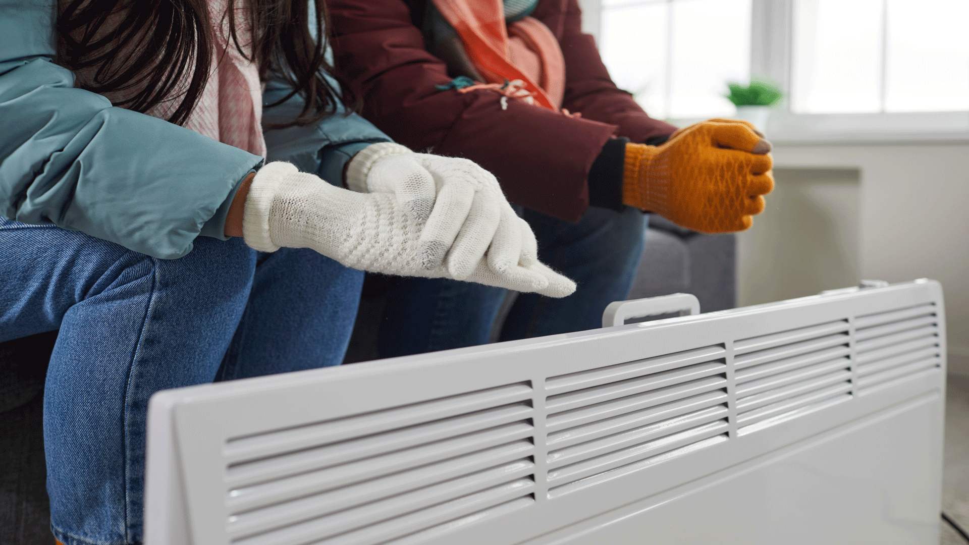 Man and woman wearing coats and gloves are rubbing their hands and warming by electric heater 