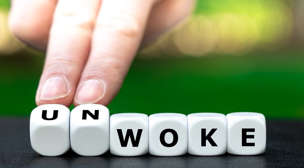 A close-up of a hand rotating a row of white dice-like cubes on a dark surface, changing the word from ‘WOKE’ to ‘UNWOKE’—the cubes spell ‘W O K E,’ and the hand is turning two cubes at the start to add ‘UN.’ The background is softly blurred in green tone